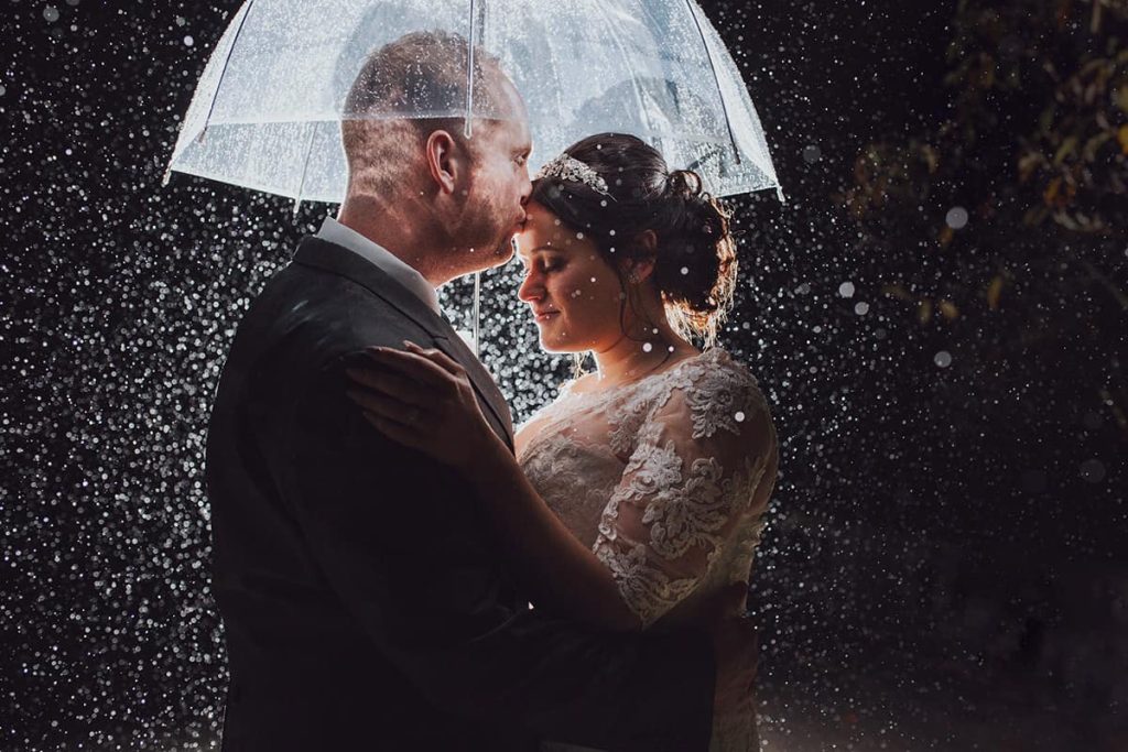 Bride and Groom stand under an umbrella at night whilst it pours with rain