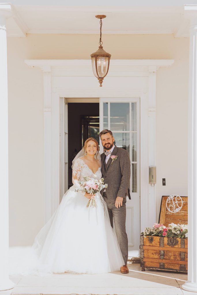 A newlywed couple stand and smile in the doorway of the manor for a photo.