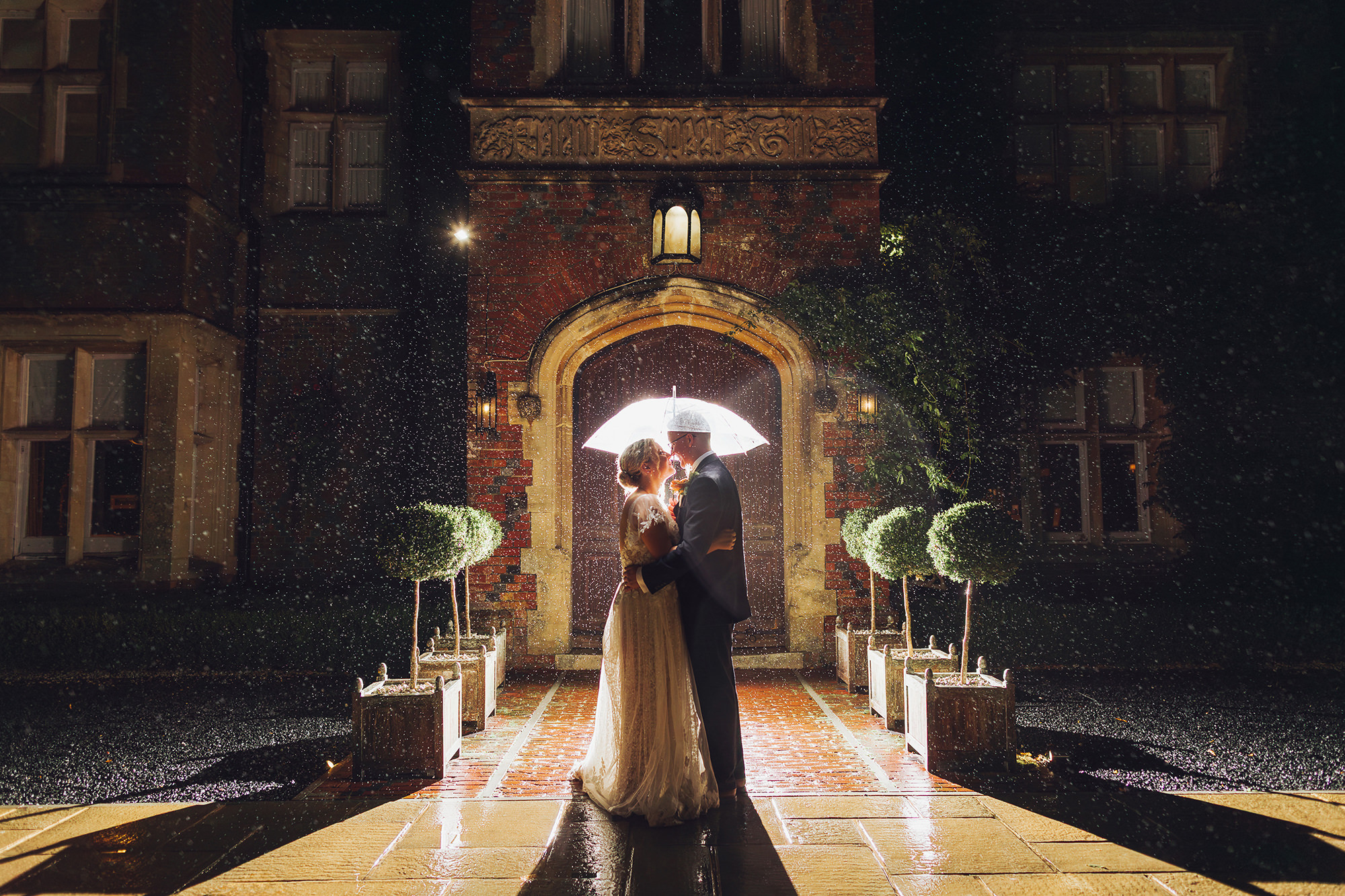 married couple stand under an umbrella outside burley manor inn the dark as the rain is lit up around them